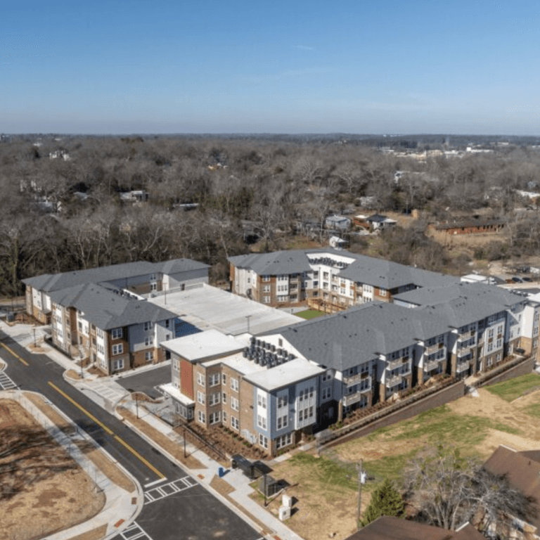 Aerial view of The View at NoDA with multiple three-story buildings, featuring a mix of brick and white panel exteriors, pitched gray roofs, and landscaped walkways. The development is surrounded by roads, green spaces, and a wooded area in the background.